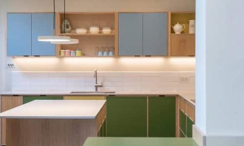 Modern kitchen with green and blue cabinets, open shelves, and a central island under a hanging light.