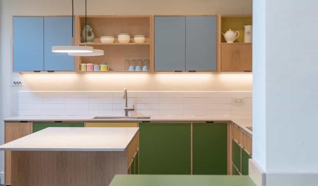 Modern kitchen with green and blue cabinets, open shelves, and a central island under a hanging light.