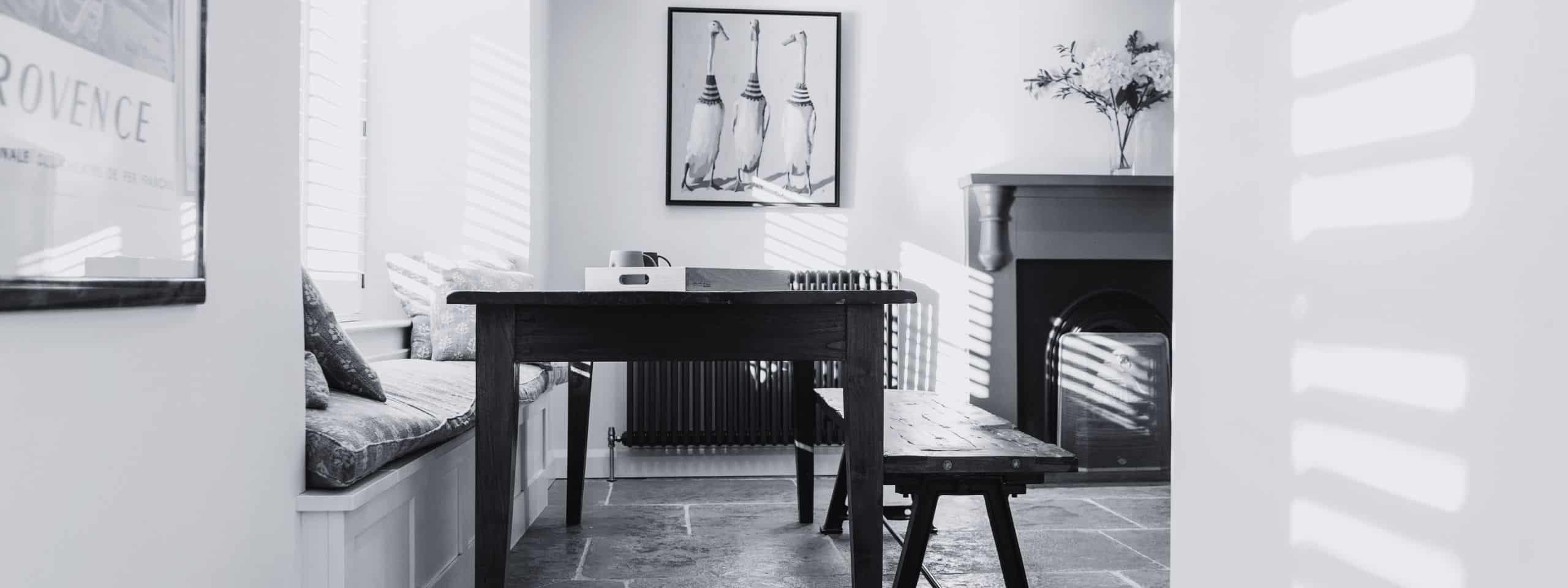 Black and white image of a cozy room with a wooden table, a bench, cushions, and wall art.
