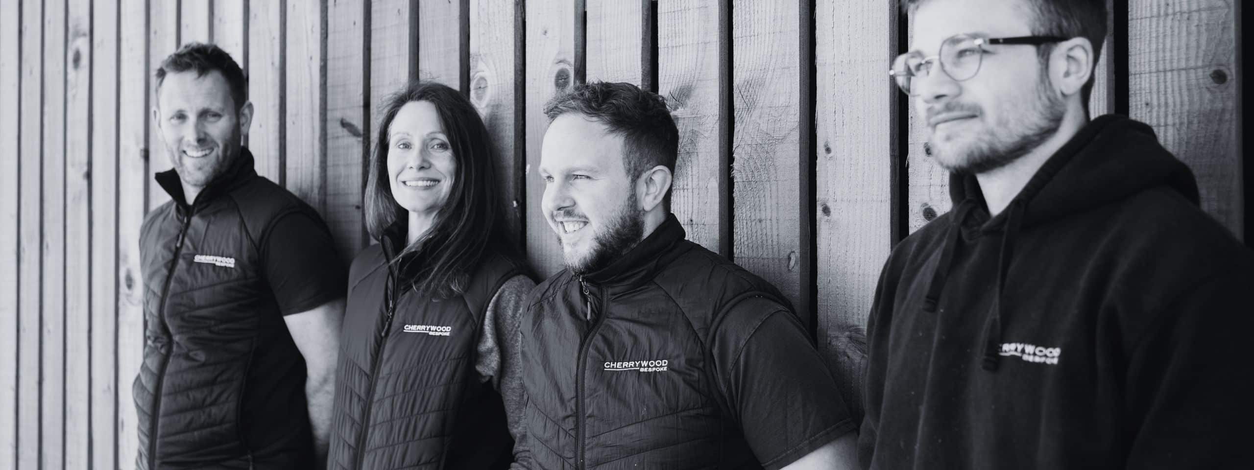 Four people in vests and hoodies standing against a wooden wall, smiling and looking off-camera.