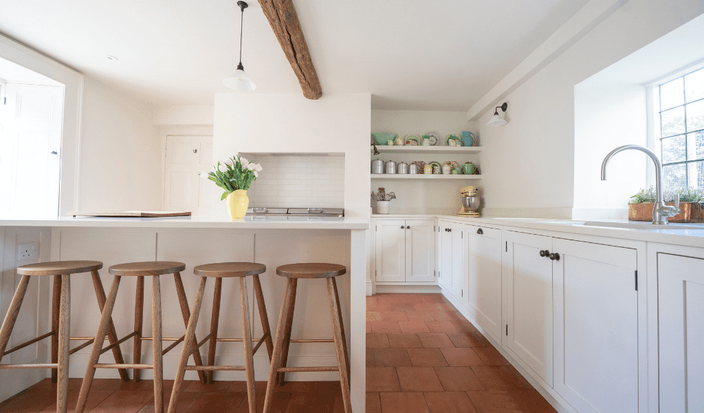 Bright kitchen with wooden stools, white cabinets, and terracotta floor. A vase with flowers is on the counter.