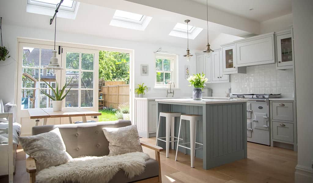 Bright kitchen with skylights, plants, and a sofa near a dining table, leading to a garden outside.