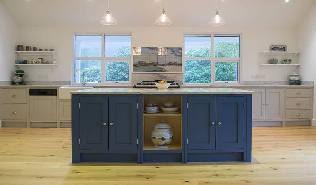 Modern kitchen with a blue island, wooden floor, and windows overlooking greenery.