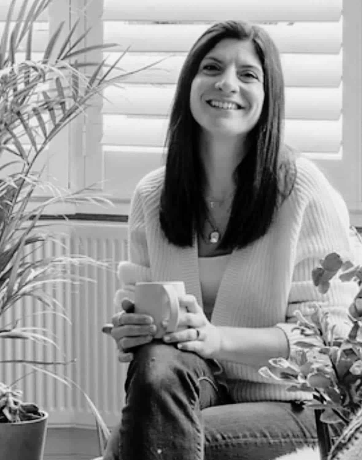 Smiling person holding a mug, surrounded by plants, sitting in front of window shutters.
