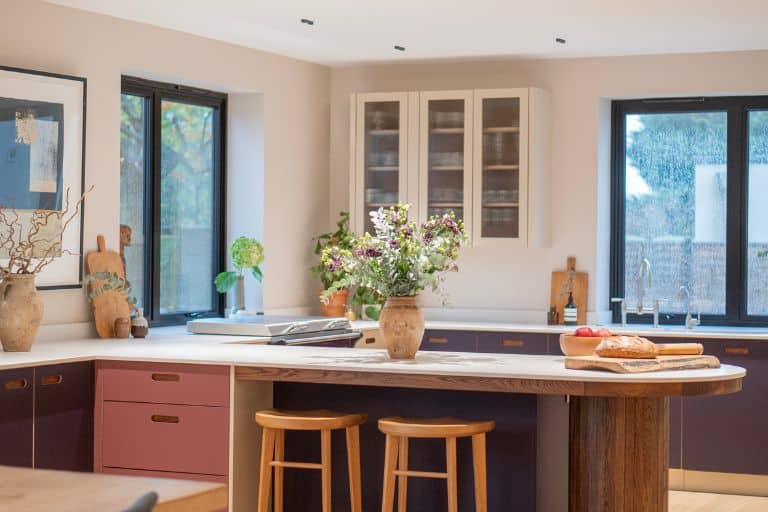 Bright kitchen with white cabinets, wooden stools, open shelves, and sunlight streaming through the windows.